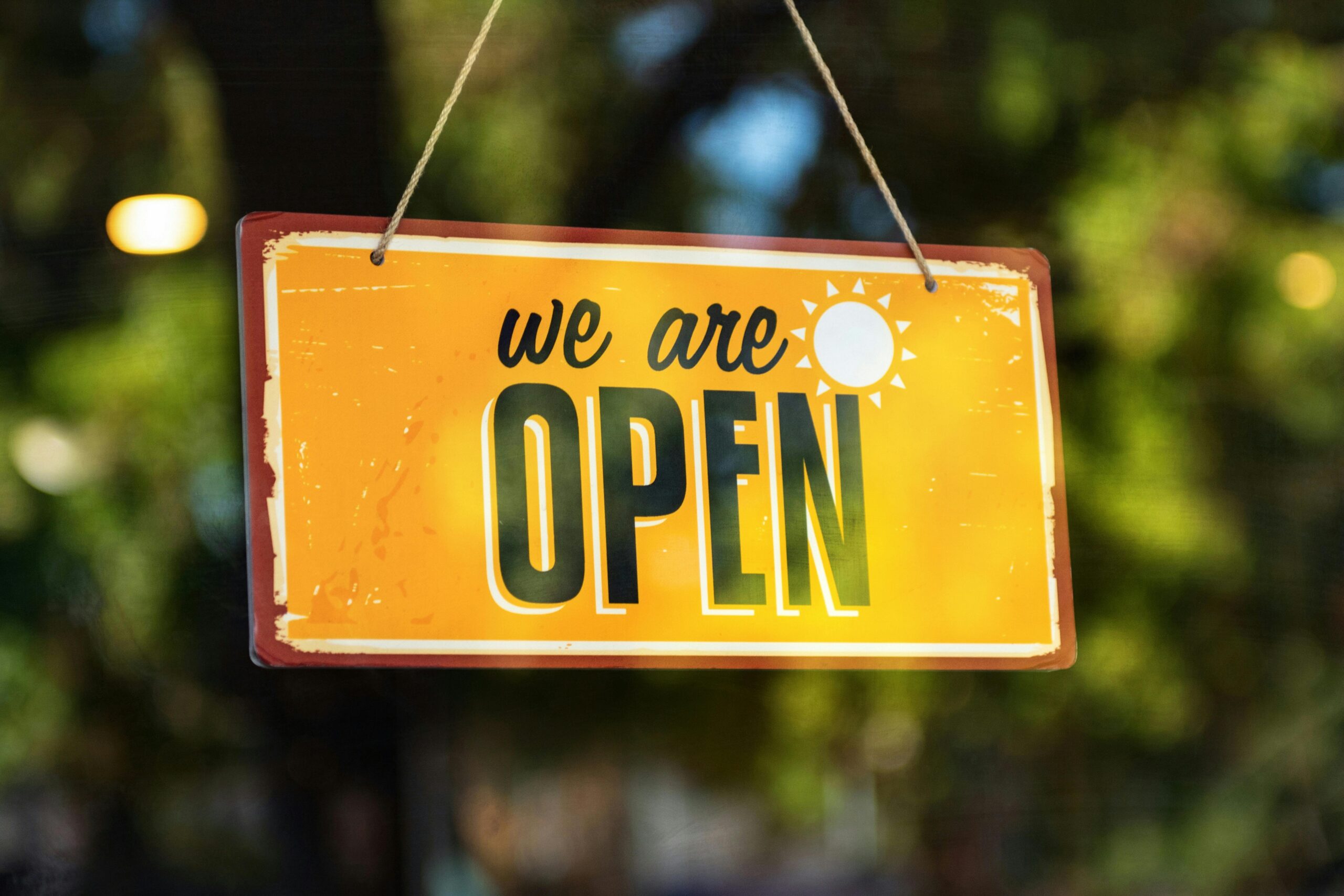 Bright yellow 'We Are Open' sign hanging on a glass door with reflections.