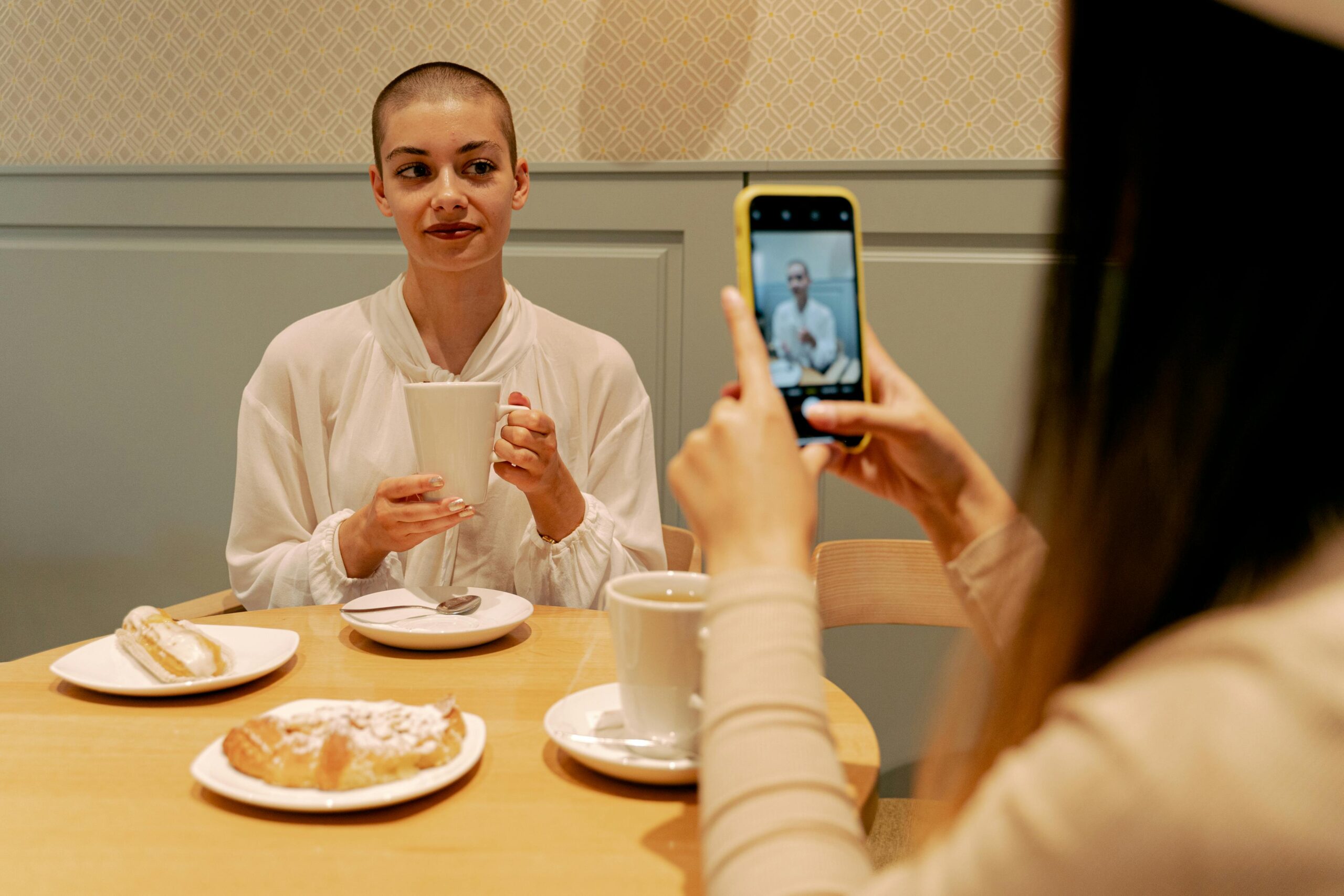 A woman with short hair enjoys coffee while a friend captures the moment. Indoor setting.