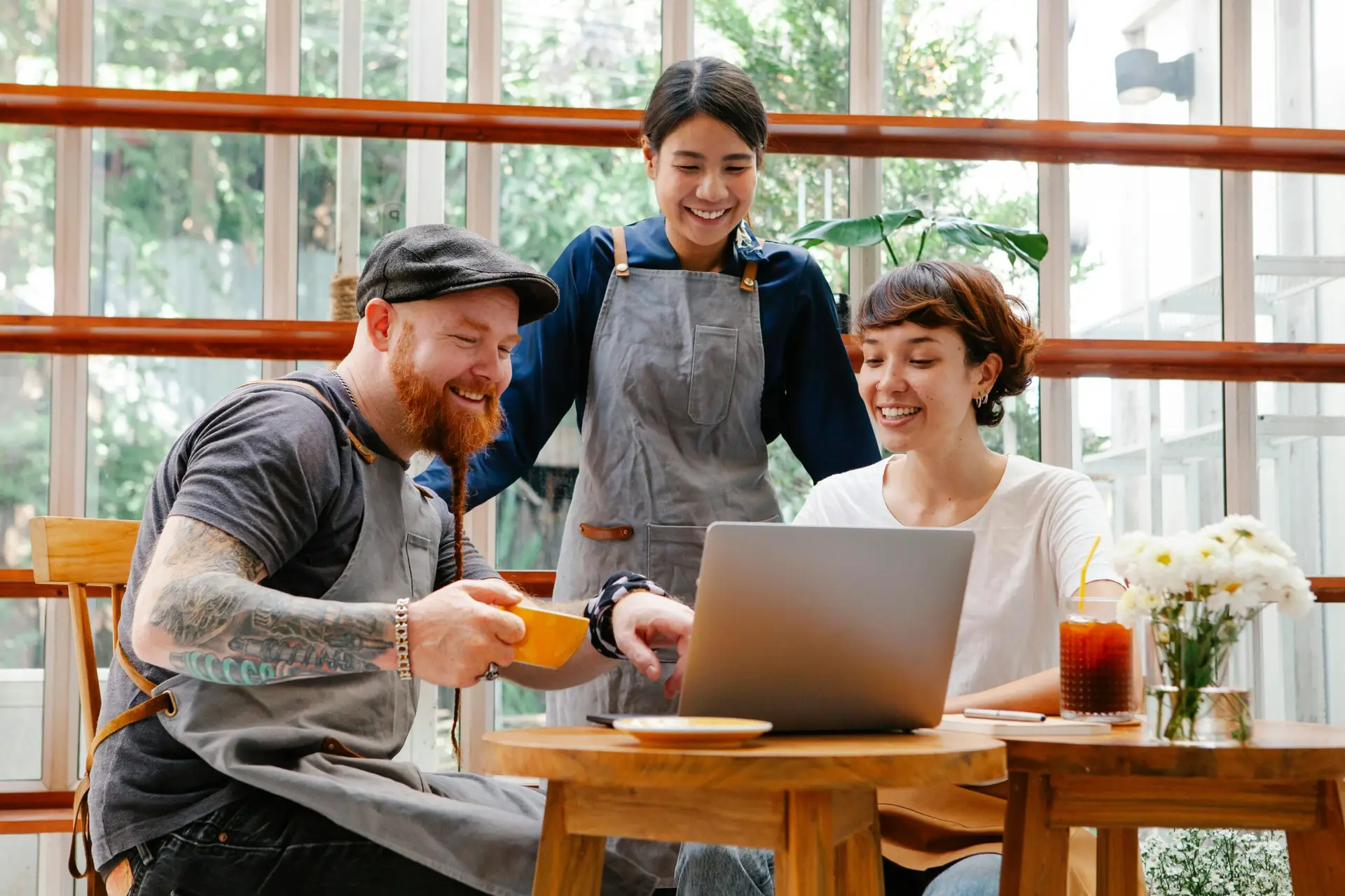 Positive coworkers in aprons and casual outfit using computer in cafeteria at table with coffee cup and glass of drink in day